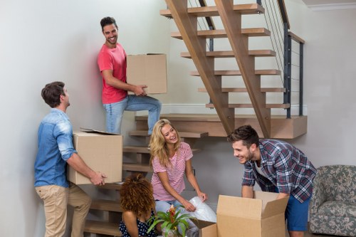 A group of five people, including three men and two women, are engaged in a house moving activity inside a modern living room. One man, dressed in a pink t-shirt and jeans, is standing on the lower staircase step holding a large cardboard box. Another man, wearing a blue shirt and beige trousers, is passing a similar box to a woman with long blonde hair, who is sitting on the floor near a small table with some items on it. The third man, in a checkered shirt, is kneeling on the floor, managing an open cardboard box. A woman with curly hair, wearing a polka-dot dress, is kneeling next to the blonde woman, possibly preparing or organizing items. The background features a contemporary staircase with wooden treads and black metal railings, leading to the upper floor, and the room has neutral-toned walls and furnishings. Natural light fills the space, emphasizing the activity of packing and moving household belongings, indicative of a house removals process in Harrow or the surrounding area, with the scene executed by Harrow Man with Van as part of their removals services.