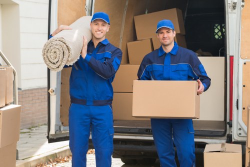 Two movers from Harrow Man with Van, dressed in blue uniforms and caps, stand outside a residential property in front of an open removals van. The first mover on the left is smiling and holding a rolled-up beige carpet, draped over his right shoulder. The second mover on the right is also smiling and holding a large cardboard box close to his chest. The van's interior visible behind them contains several brown cardboard boxes stacked neatly, with one positioned on top of another, ready for transportation. The scene takes place on a residential street, with a partially visible house wall and garden area. The lighting appears natural, suggesting daytime conditions. The image reflects a professional house removals service in a suburban area, with focus on the preparation and careful handling of household items, including soft furnishings and boxes, typical of removals in Harrow or nearby areas within the HA postcode of Greater London.