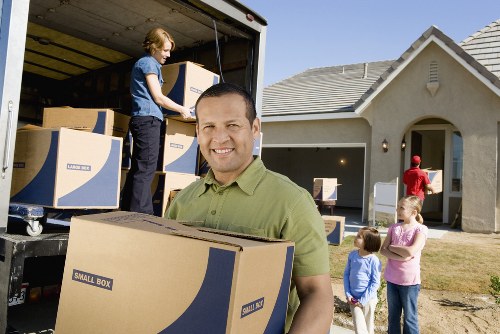 A professional house removal scene showing three individuals involved in a relocation on a residential street in Harrow, near the postcode area. In the foreground, a man with short, dark hair and a green polo shirt is smiling and holding a medium-sized cardboard box labeled 'Small Box,' prepared for moving. Behind him, a woman with blonde hair, dressed in a blue top and dark trousers, is standing inside the open back of a black removals van, lifting and handling additional plain cardboard boxes. To the right, a man in a red shirt is seen near the entrance of a modern detached house with a tiled roof, light-colored exterior walls, and a small front garden. Two young children, one in a pink top and another in a blue outfit, stand nearby watching the activity, indicating a family moving process. The setting appears to be a sunny day, with clear skies and well-maintained grass on the lawn, contributing to a typical suburban environment consistent with house removals in the Harrow area, as managed by Harrow Man with Van. The scene captures the essentials of packing, lifting, and loading furniture and boxes, showcasing a professional removal service in action on a residential street.