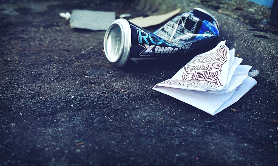 A close-up photograph of an empty, crushed energy drink can lying on a paved surface outdoors, with a crumpled sheet of paper containing printed diagrams or hieroglyphic-like symbols near it. The surf
