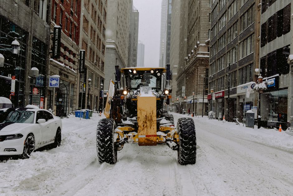 A heavy-duty snow plough attachment mounted on the front of a large vehicle, likely a tractor or a large van, actively clearing snow from a roadway during winter. The snow, which appears compacted and
